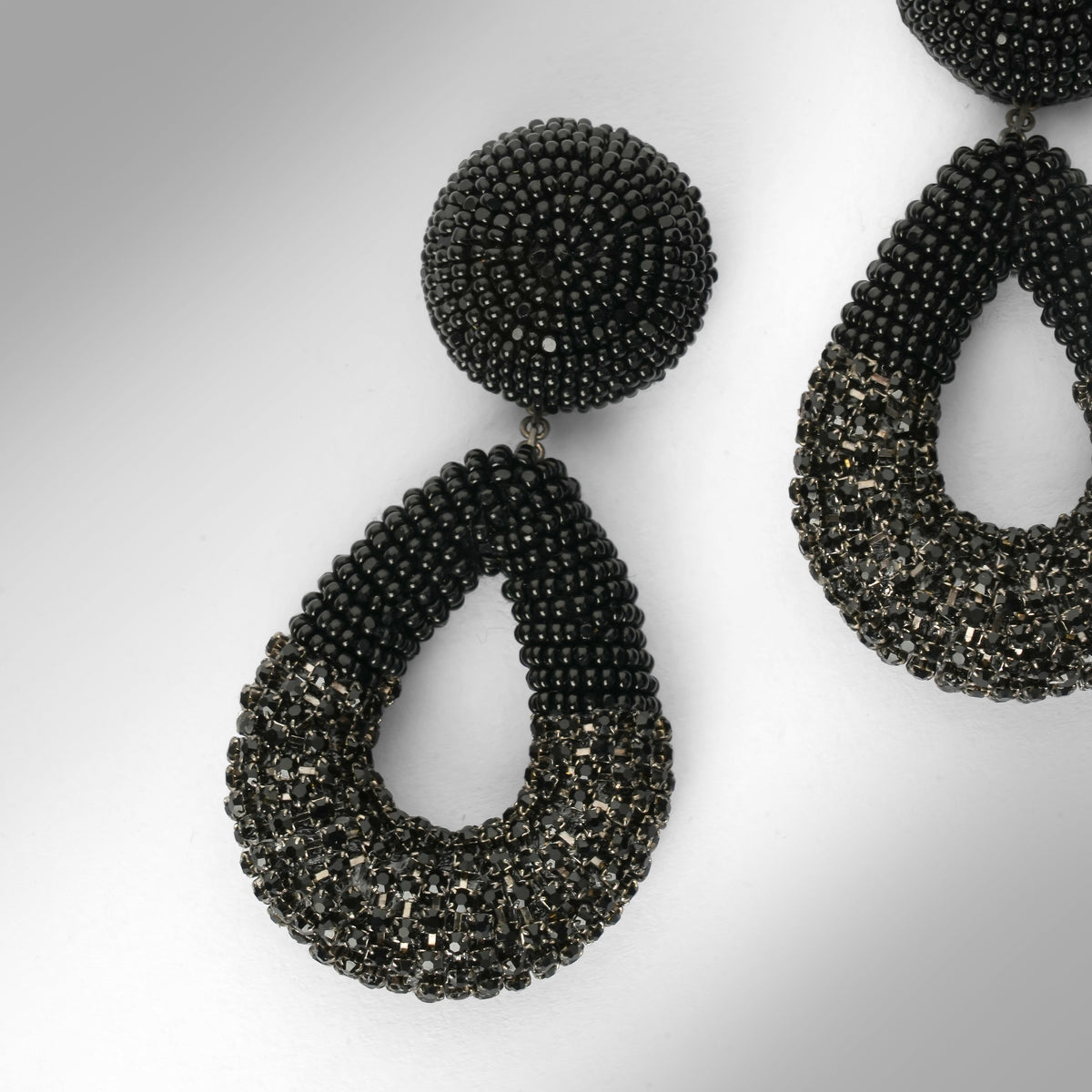 Close up of A pair of large statement dangle earrings made from black beads and chainstone, featuring a round stud top and a teardrop-shaped drop, shown on a light gray gradient background.