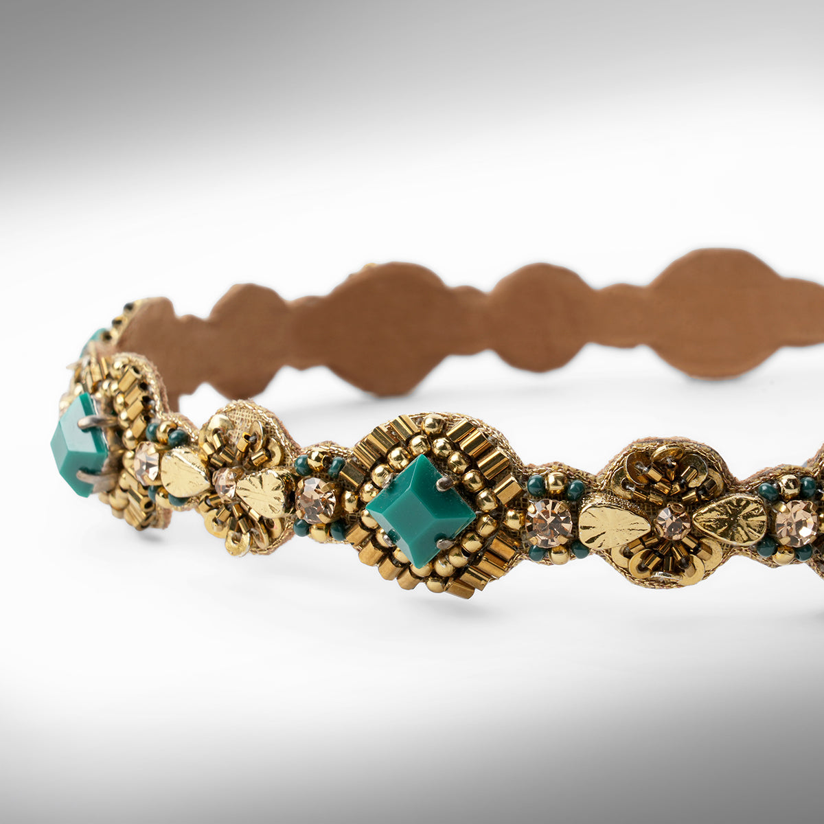Close up of an ornate gold-toned headband with emerald stone accents shown against a neutral gray background.