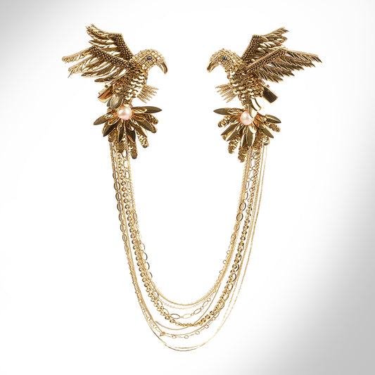 A gold-toned brooch featuring detailed bird motifs with multiple dangling chain layers, displayed on a grey background.
