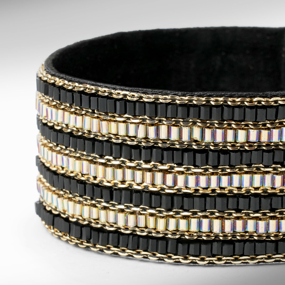 A close up of a wide beaded cuff bracelet featuring black faceted beads, gold chain links, and iridescent beads set against a neutral gray background.
