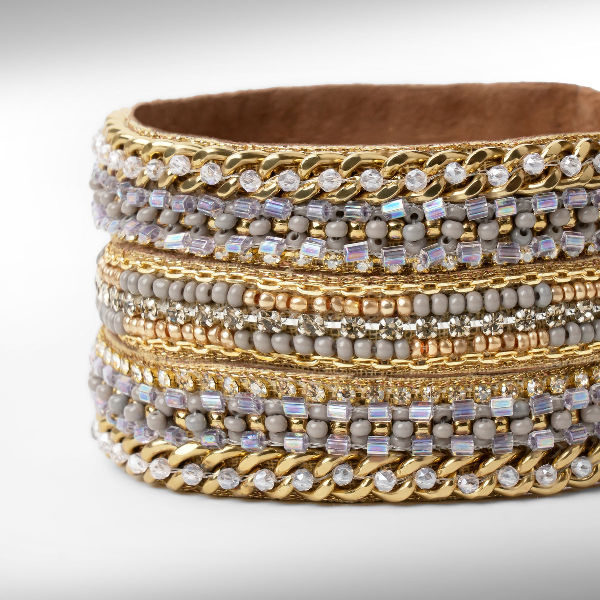 A close-up of a gold-tone bracelet with mixed textures including chain links, beads, and clear stones, set against a light gray background.