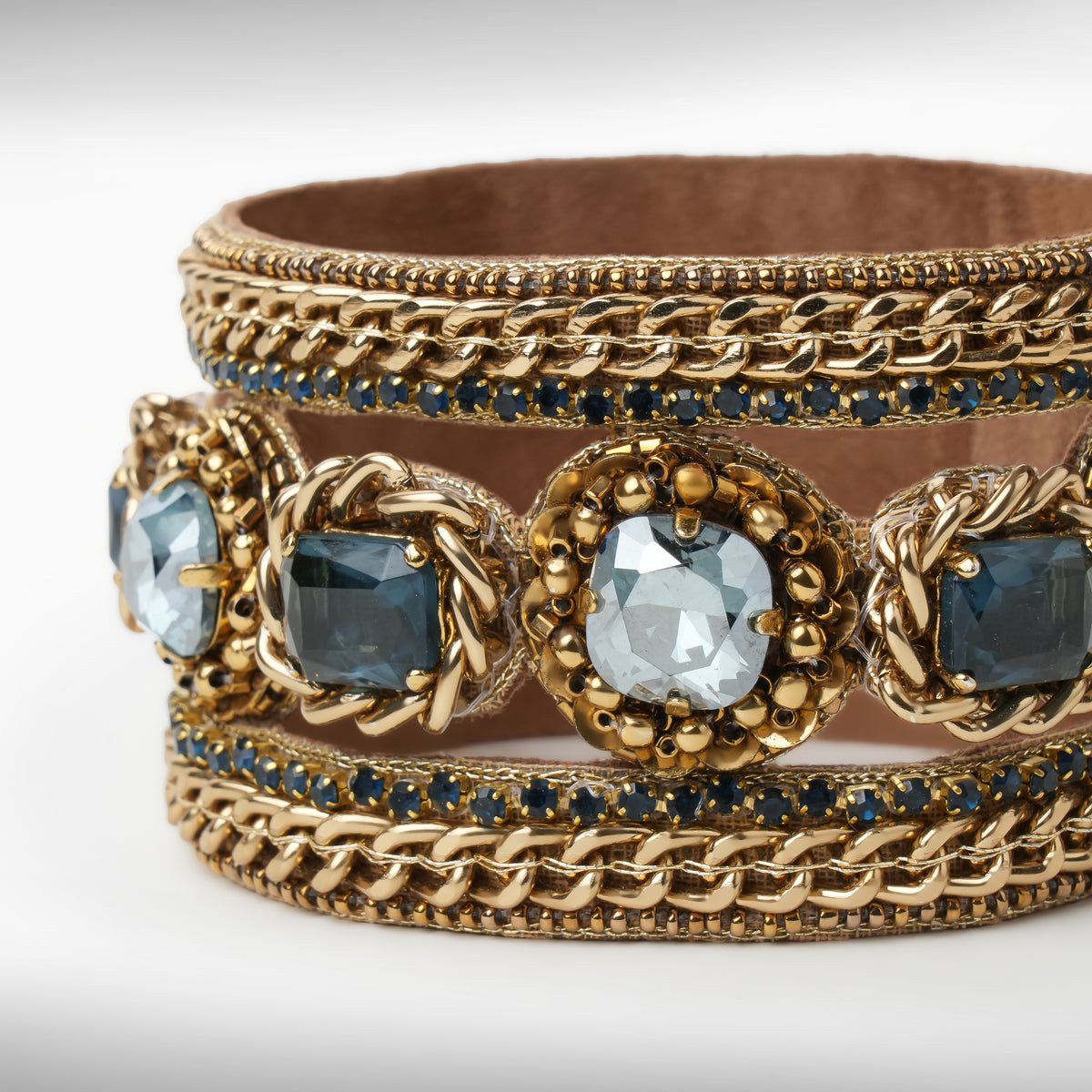 Close up of A wide gold-tone cuff bracelet featuring woven chain details, ornate floral medallions with blue and sapphire faceted crystals, and rows of small sapphire stones, shown on a light gray background.