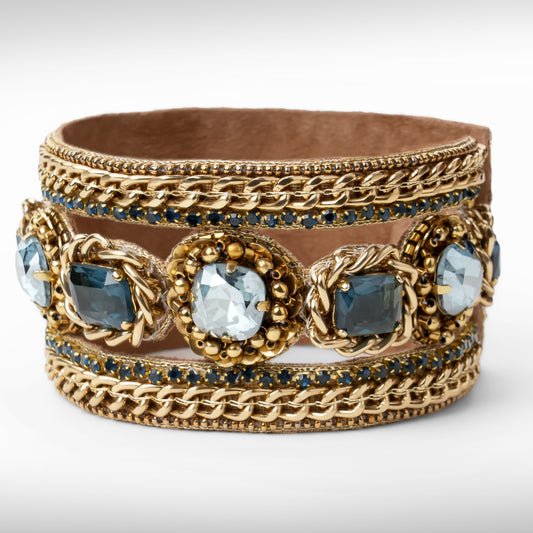 A wide gold-tone cuff bracelet featuring woven chain details, ornate floral medallions with blue and sapphire faceted crystals, and rows of small amethyst stones, shown on a light gray background.