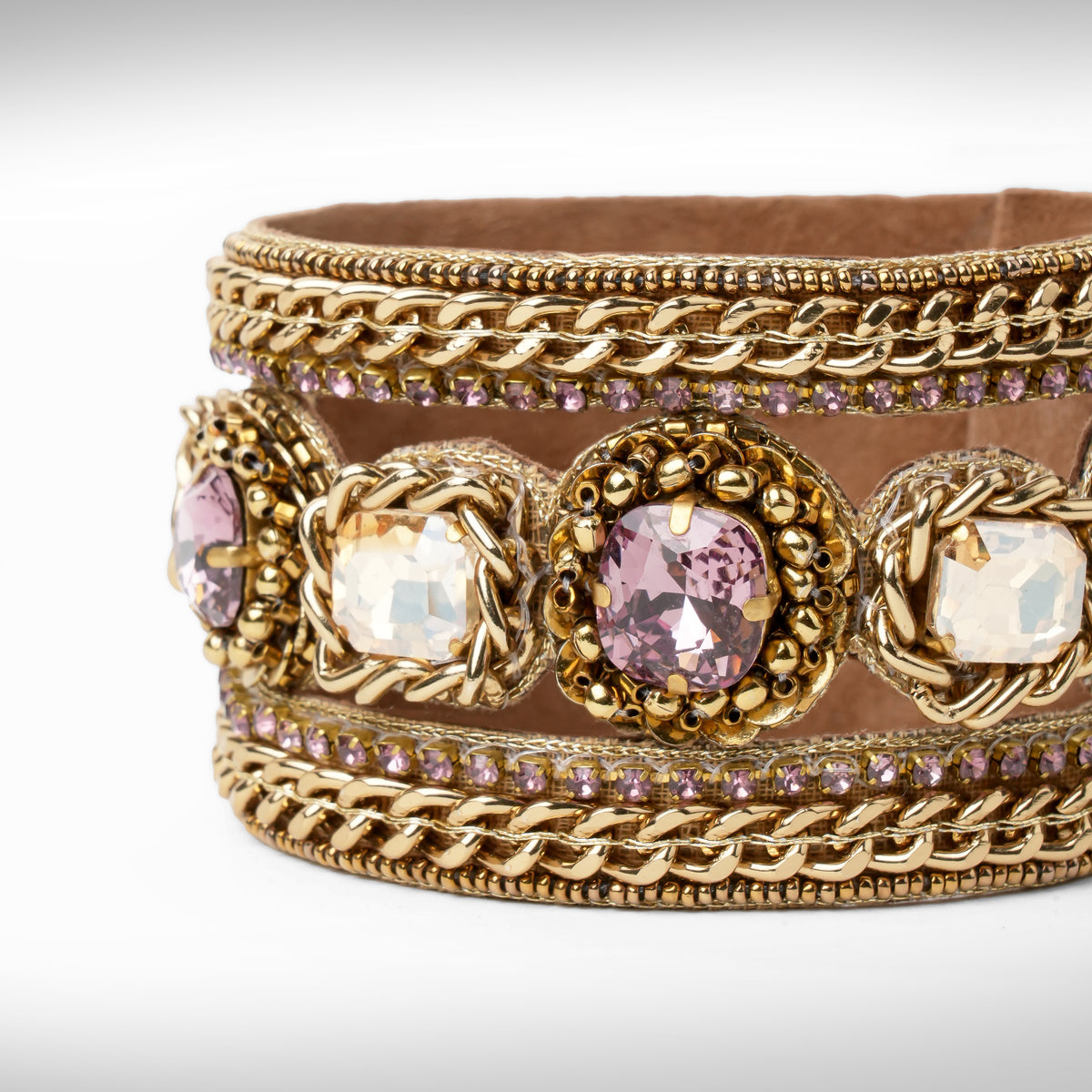 Close up of A wide gold-tone cuff bracelet featuring woven chain details, ornate floral medallions with clear and amethyst faceted crystals, and rows of small amethyst stones, shown on a light gray background.