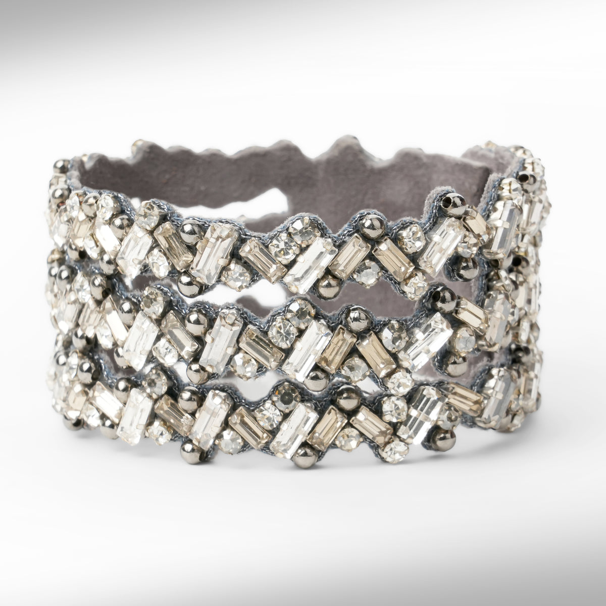a wide gold-tone cuff bracelet embellished with multiple rows of gunmetal and silver-toned rectangular and round crystals, set against a plain white background.