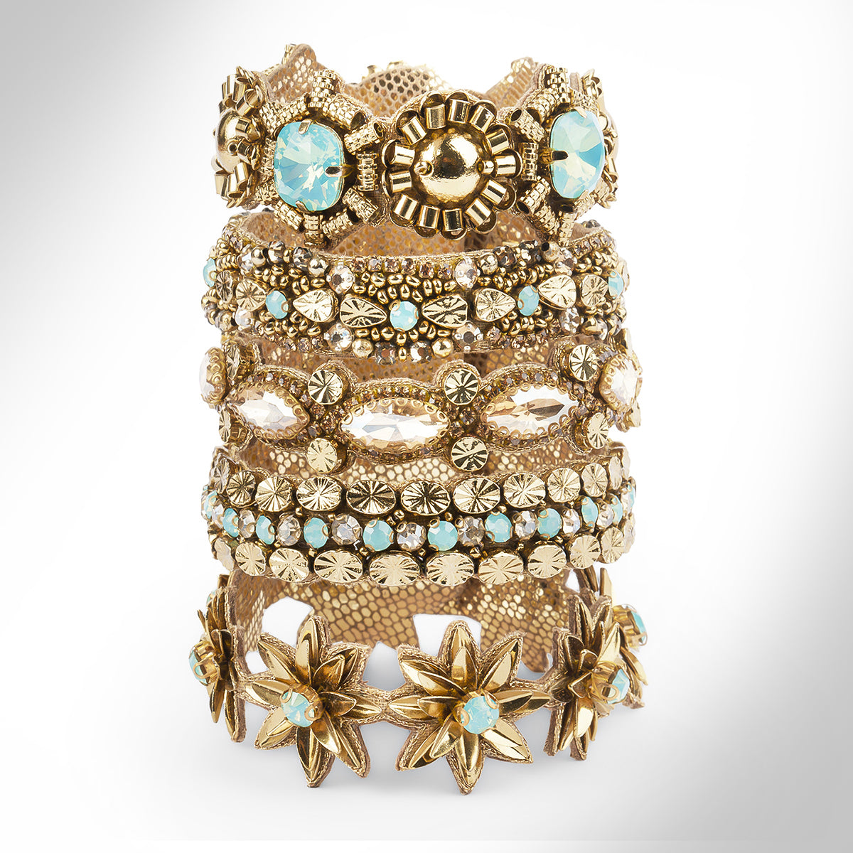 A wide, multi-layered gold-toned bracelet featuring intricate floral and starburst motifs, embellished with light blue faceted stones and small turquoise-like beads, set against a neutral light gray background.