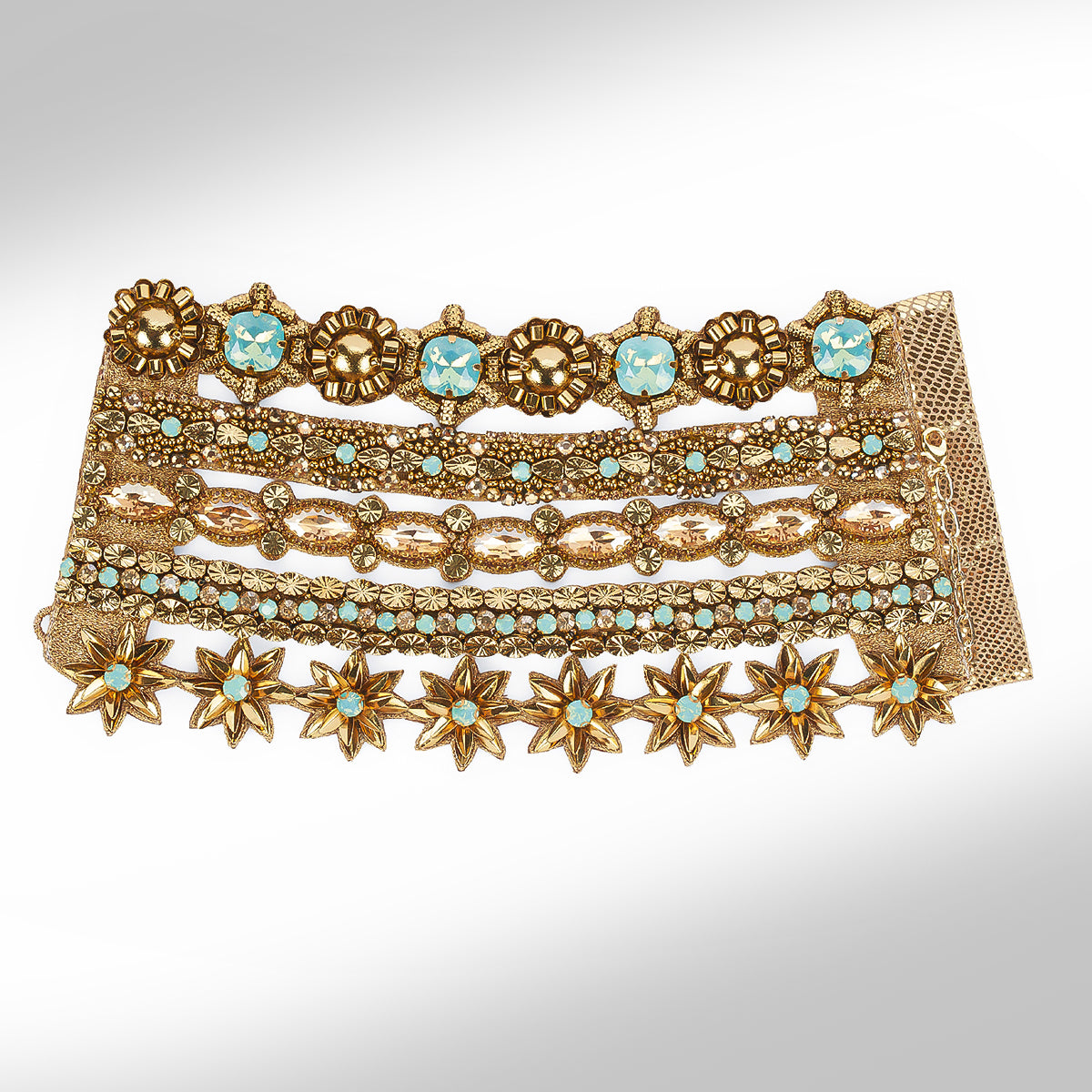 A wide, multi-layered gold-toned bracelet featuring intricate floral and starburst motifs, embellished with light blue faceted stones and small turquoise-like beads, set against a neutral light gray background.