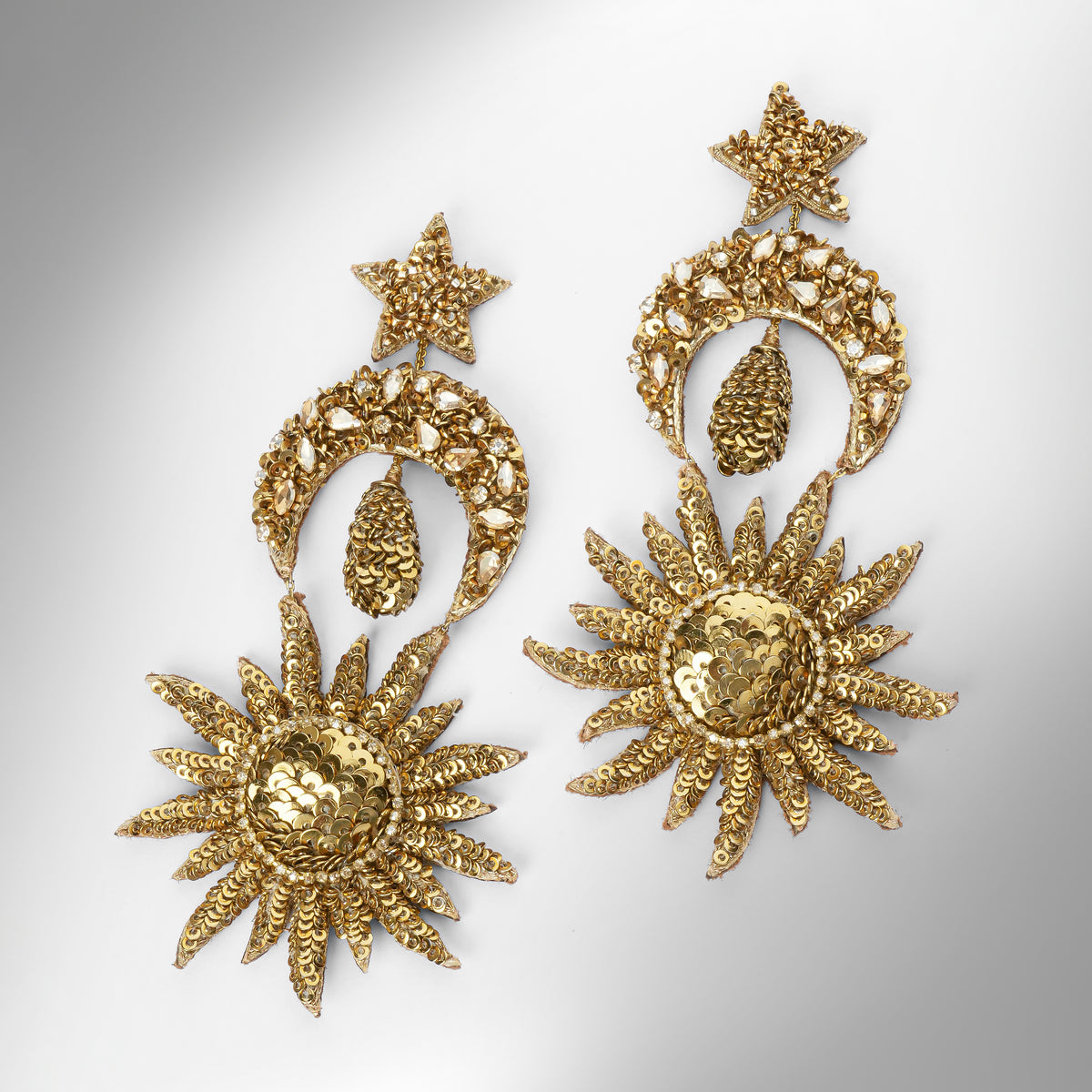 A pair of ornate gold-toned statement earrings featuring star-shaped studs, crescent moon motifs with teardrop pendants, and large sunburst drops with textured detailing, shown on a light gray gradient background.