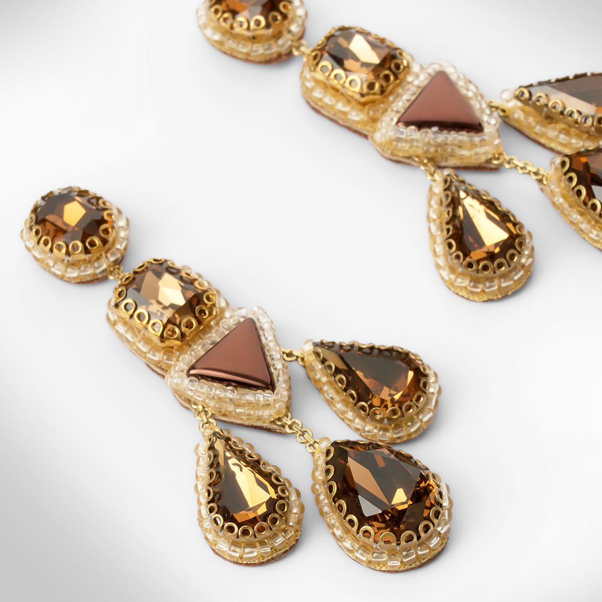 Close up of a pair of ornate gold-toned statement earrings with faceted brown and amber gemstones, displayed on a clean light gray background.