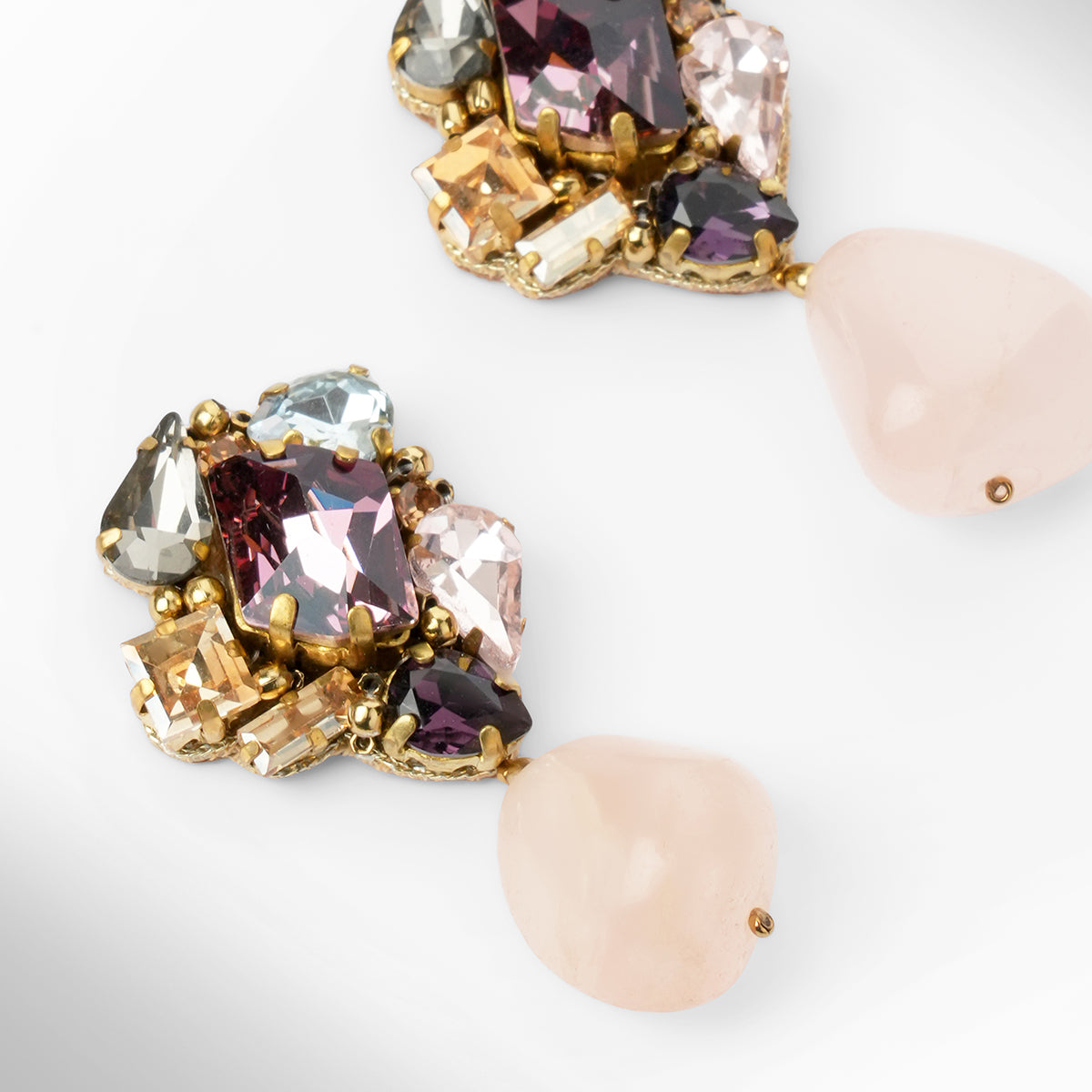 Close up of a  pair of gold-tone drop earrings featuring a cluster of multicolored faceted crystals at the top and a teardrop-shaped pale pink stone dangling below each earring, shown on a light gray background.