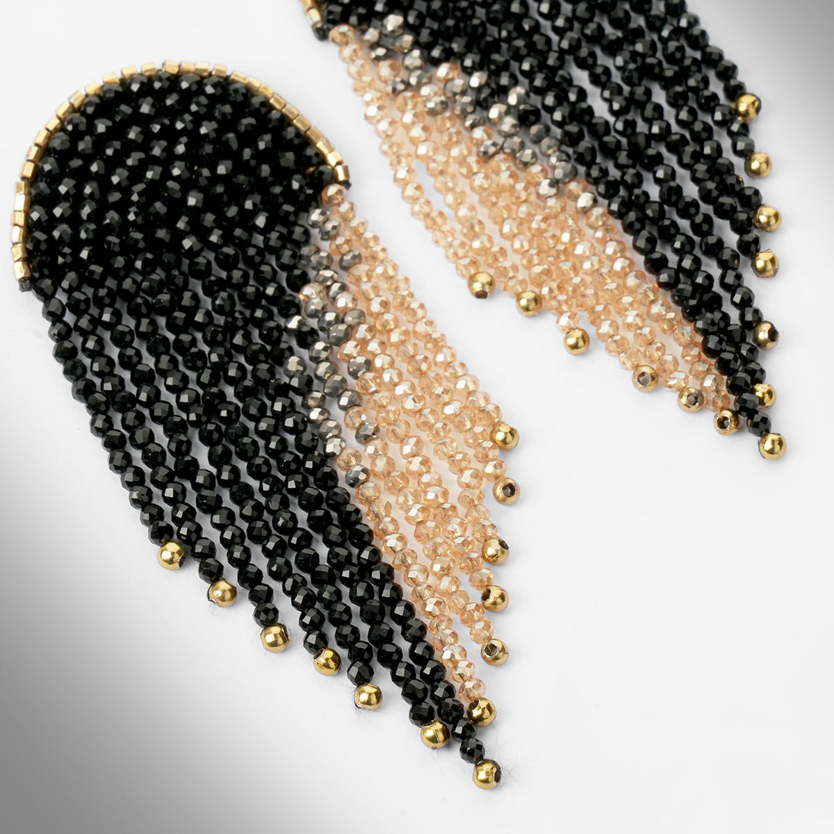 Close up of a pair of beaded fringe earrings with a black top section, transitioning into pale champagne beads, finished with small gold beads at the tips, shown on a light gray gradient background.