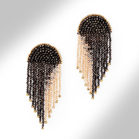 A pair of beaded fringe earrings with a black top section, transitioning into pale champagne beads, finished with small gold beads at the tips, shown on a light gray gradient background.