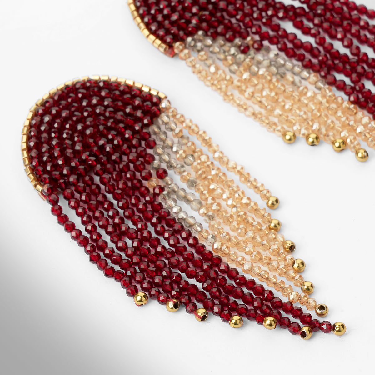 Close up of a pair of beaded fringe earrings with a dark red top section, transitioning into pale champagne beads, finished with small gold beads at the tips, shown on a light gray gradient background.