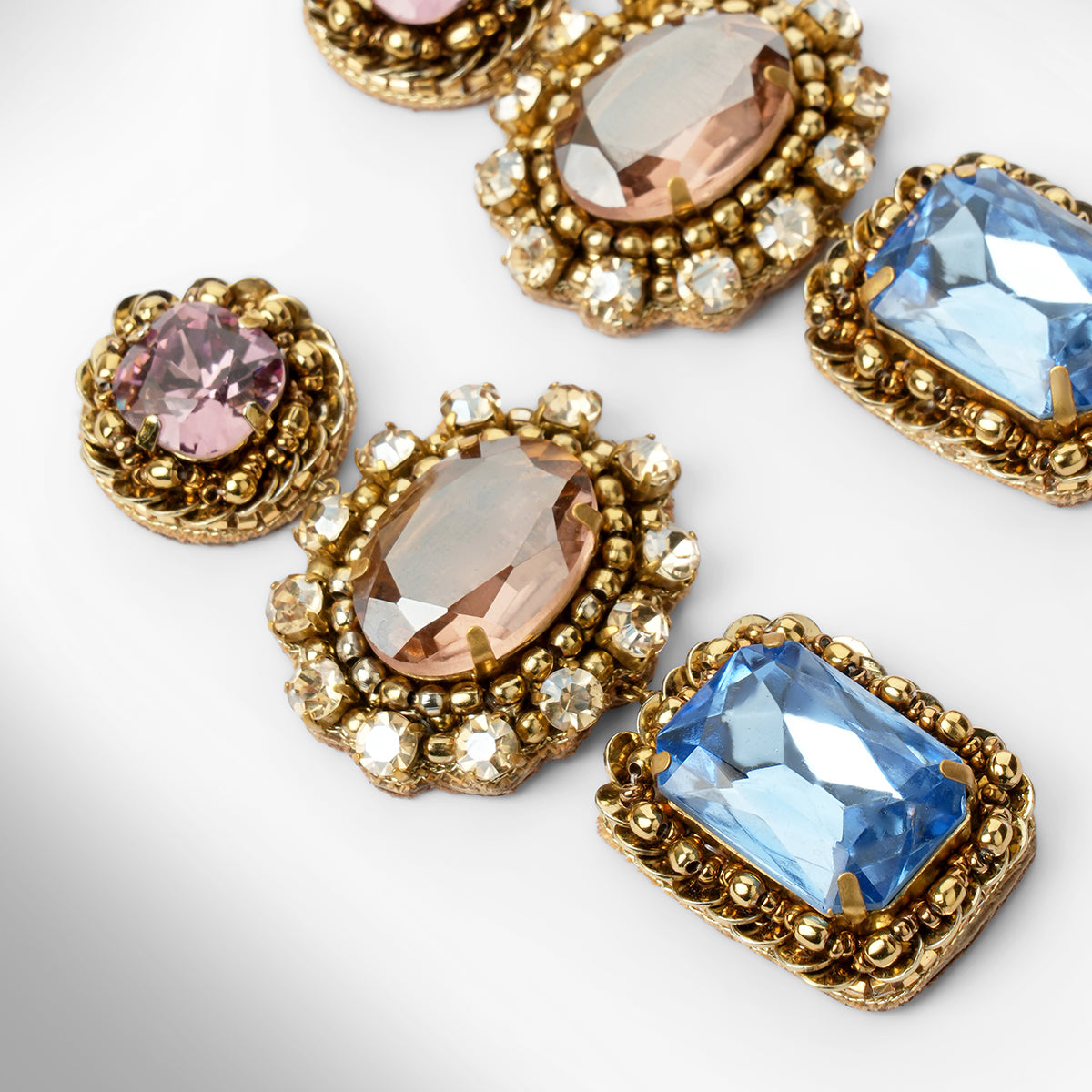 Close up of a pair of ornate gold-tone drop earrings with pink, peach, and light blue faceted stones surrounded by champagne stones, shown on a light gray background.