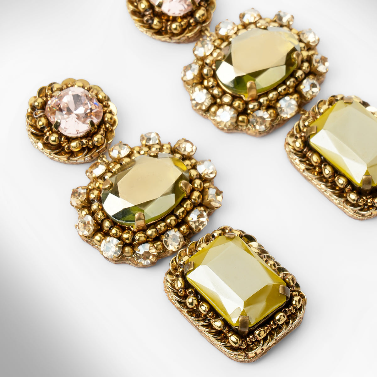 Close up of a pair of ornate gold-tone drop earrings with pink, green, and olive faceted stones surrounded by champagne stones, shown on a light gray background.