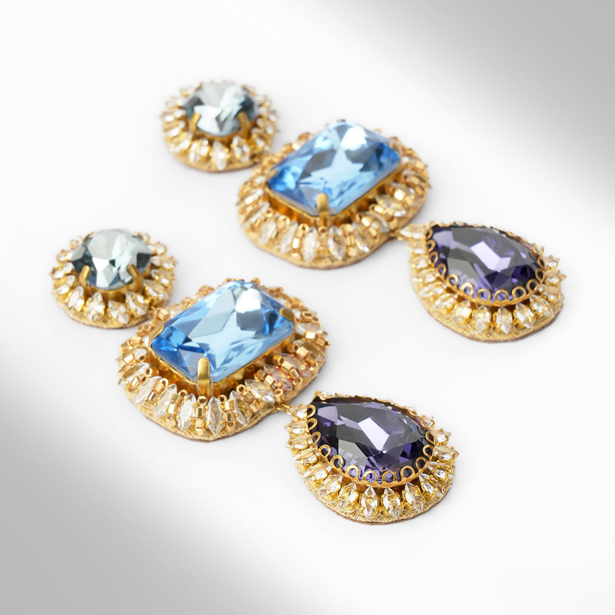 Close up of A pair of ornate drop earrings with blue and sapphire stones surrounded by champagne stones on a light gray background.