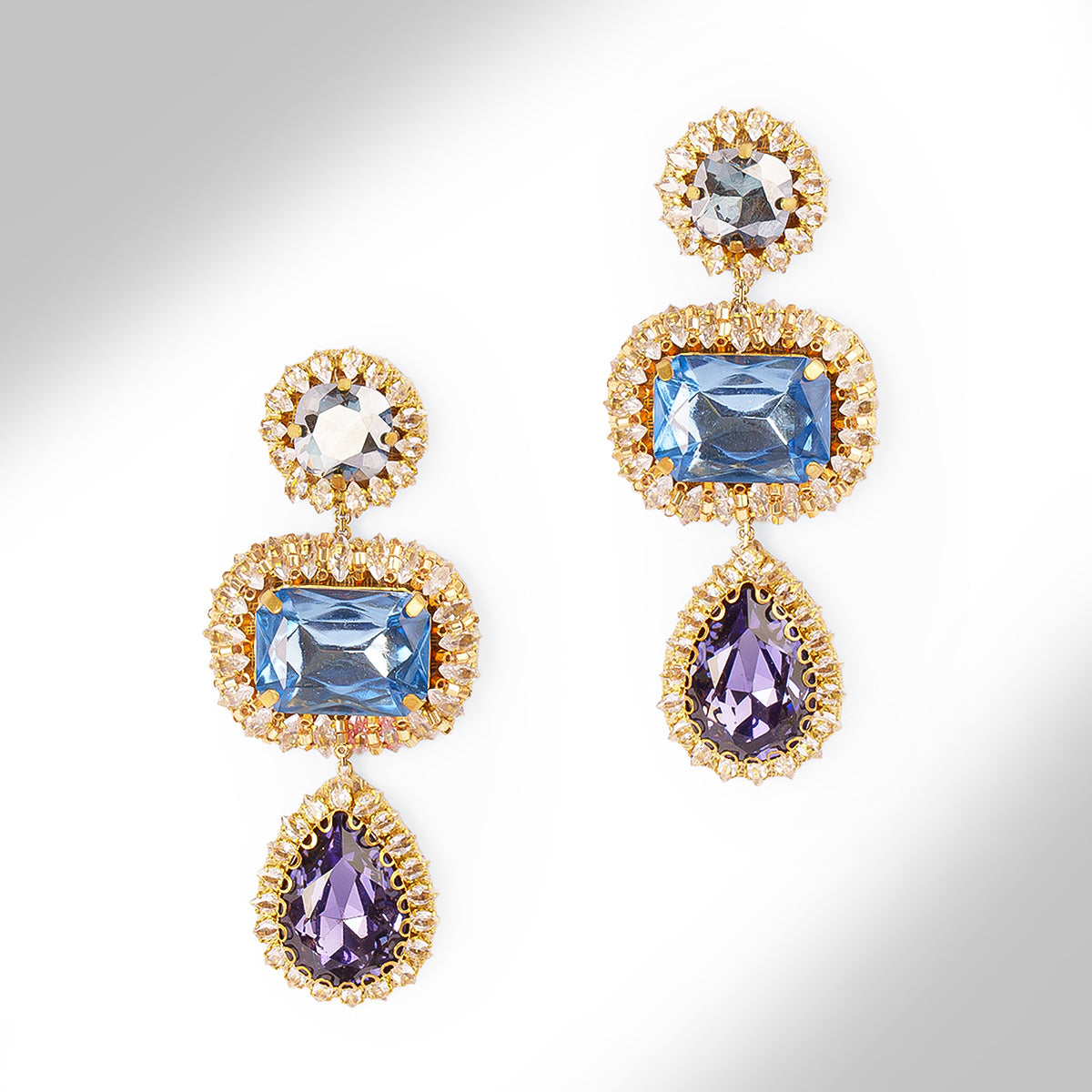 A pair of ornate drop earrings with blue and sapphire stones surrounded by champagne stones on a light gray background.