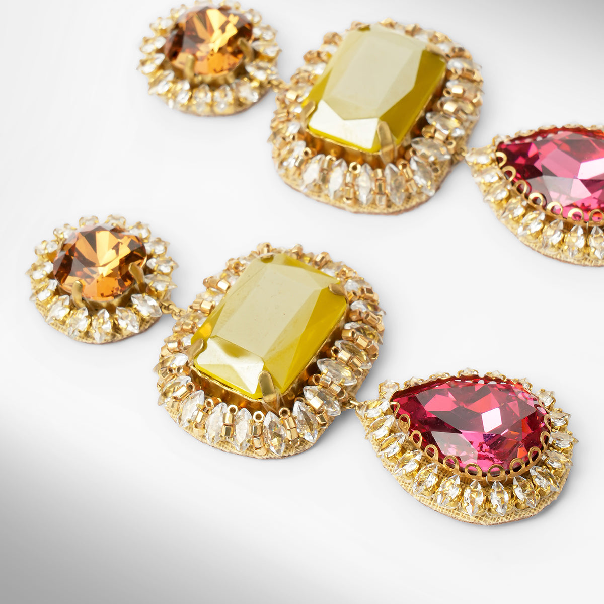 Close up of a pair of ornate drop earrings with olive and pink stones surrounded by clear stones on a light gray background.