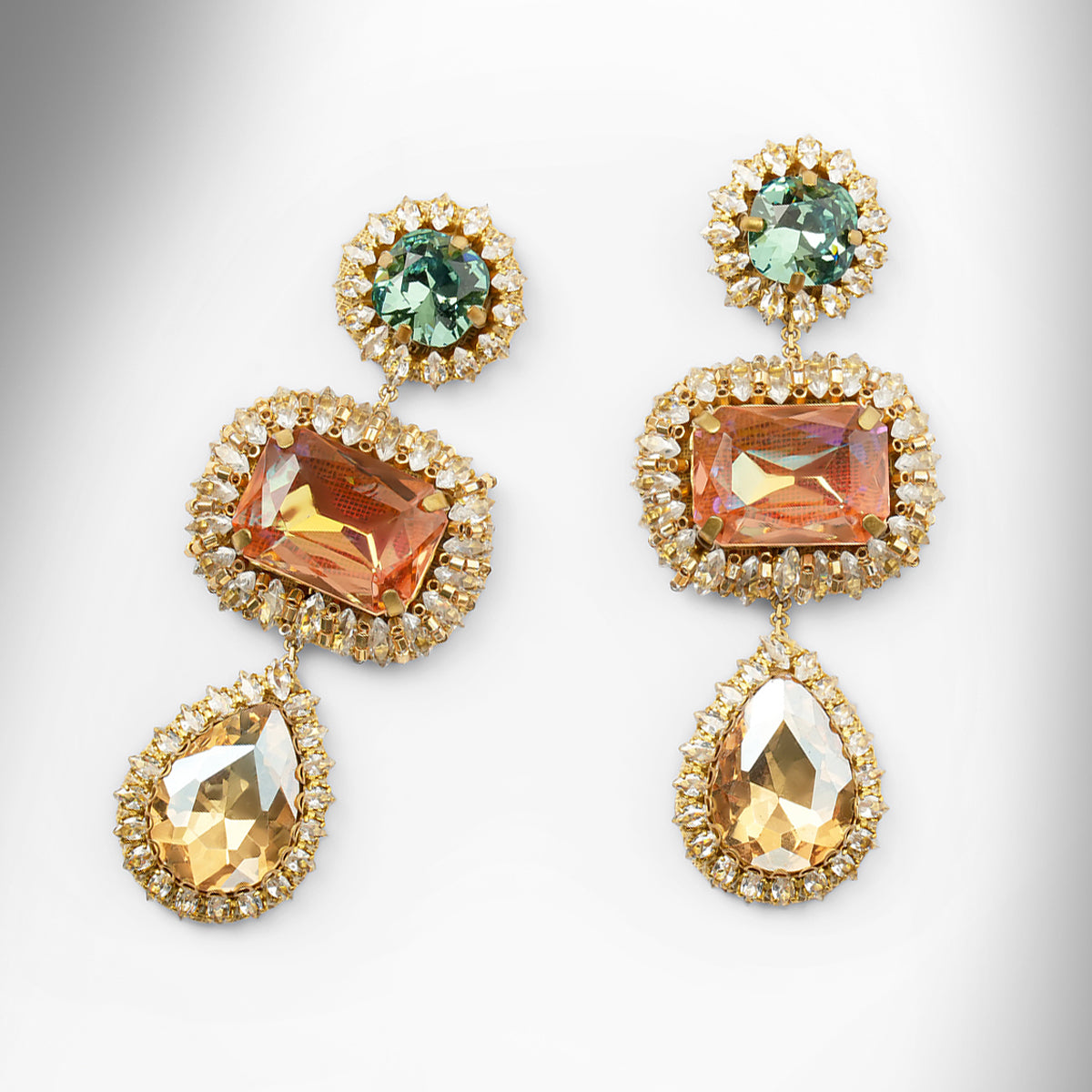 A pair of ornate drop earrings with blue and peach stones surrounded by champagne stones on a light gray background.