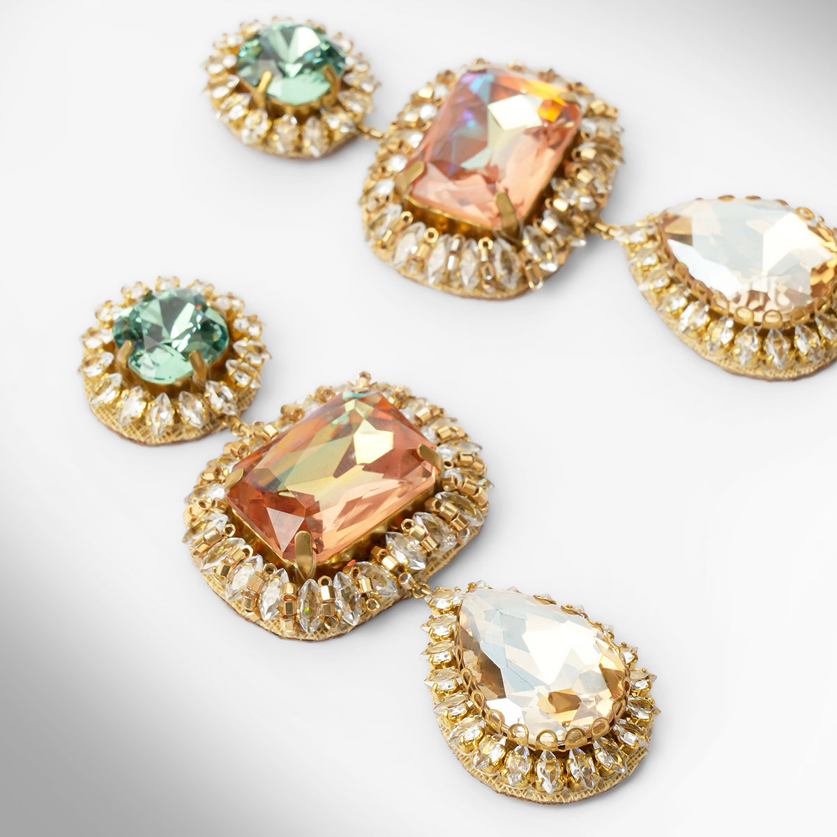 Close up of a pair of ornate drop earrings with blue and peach stones surrounded by champagne stones on a light gray background.