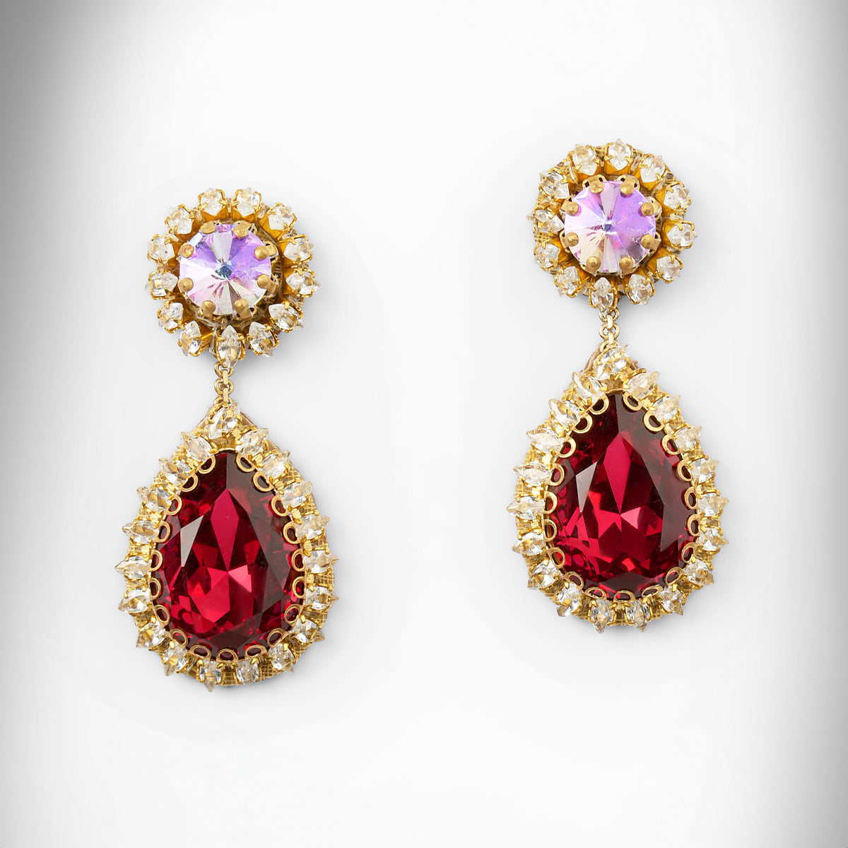 A pair of gold tone drop earrings featuring an iridescent top and a larger teardrop-shaped deep red stone below, surrounded by small clear stones.