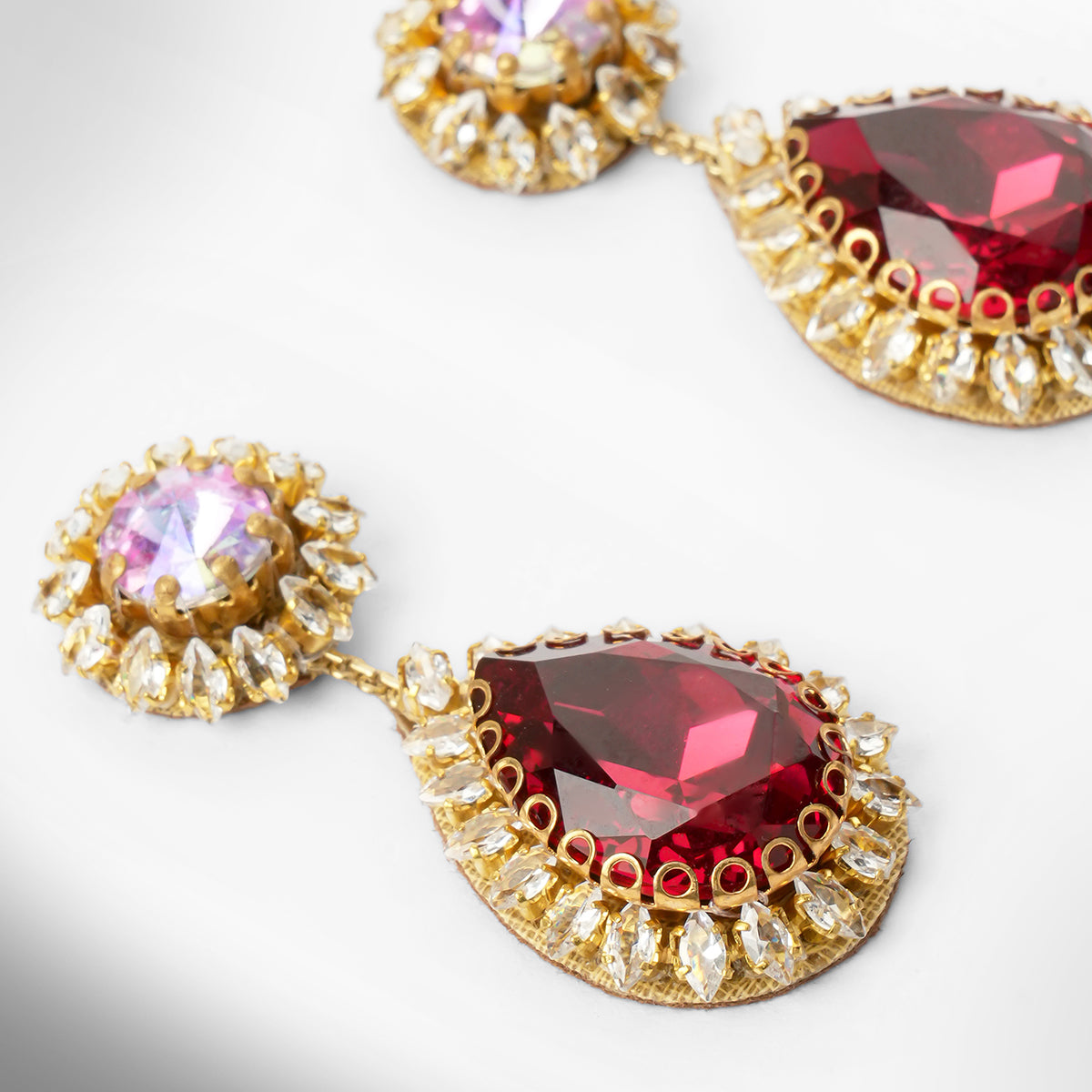 Close up of a pair of gold tone drop earrings featuring an iridescent top and a larger teardrop-shaped deep red stone below, surrounded by small clear stones.