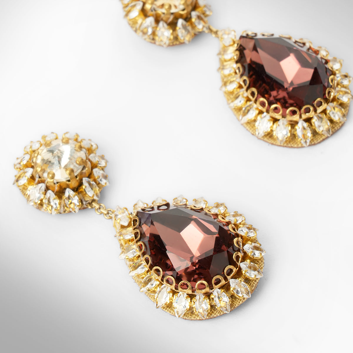 Close up of A pair of gold tone drop earrings featuring a champagne top and a larger teardrop-shaped brown stone below, surrounded by small clear stones.