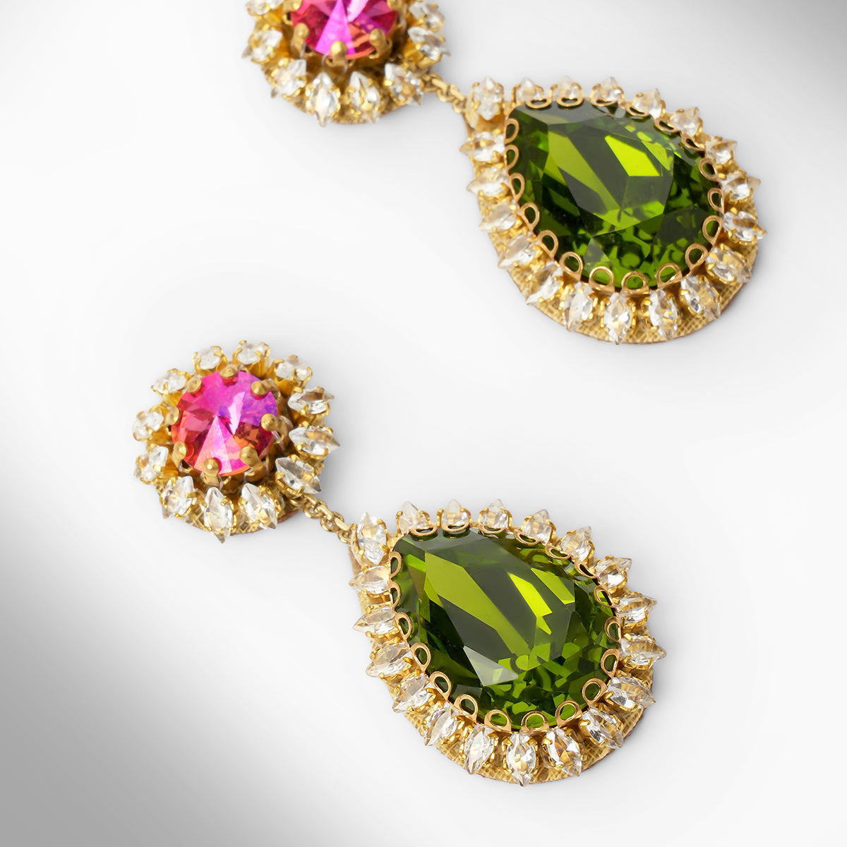 Close up of a pair of gold tone drop earrings featuring a fuchsia top and a larger teardrop-shaped green stone below, surrounded by small clear stones.