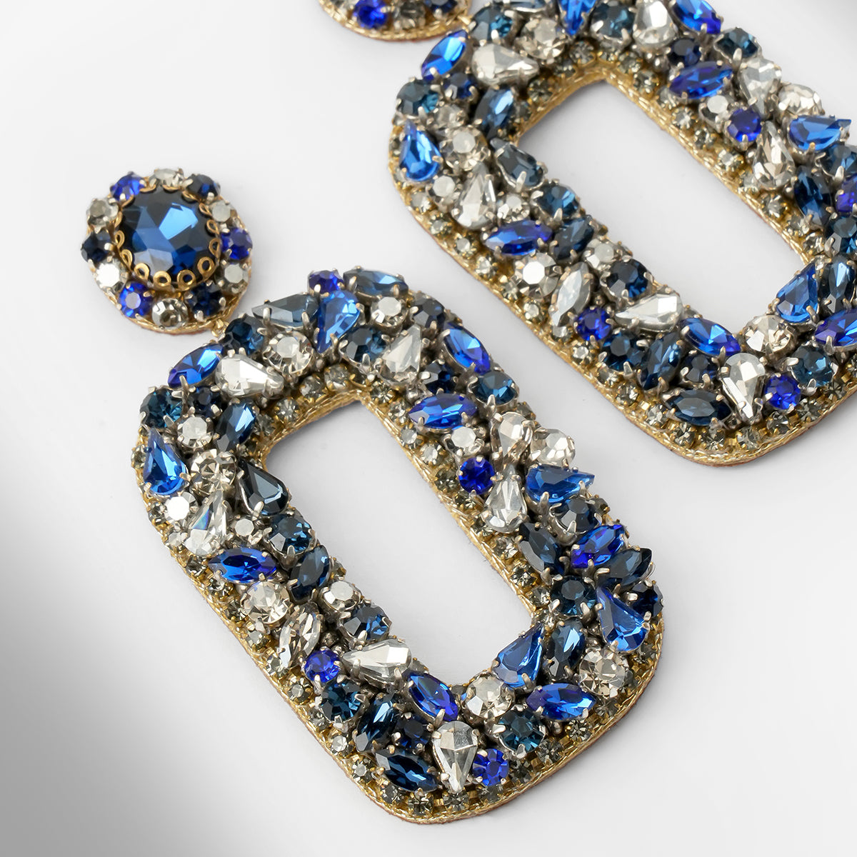 Close up of Gold-tone statement earrings with large open rectangular shapes and a matching round stud, heavily embellished with blue, silver, and clear faceted stones on a light gray background.