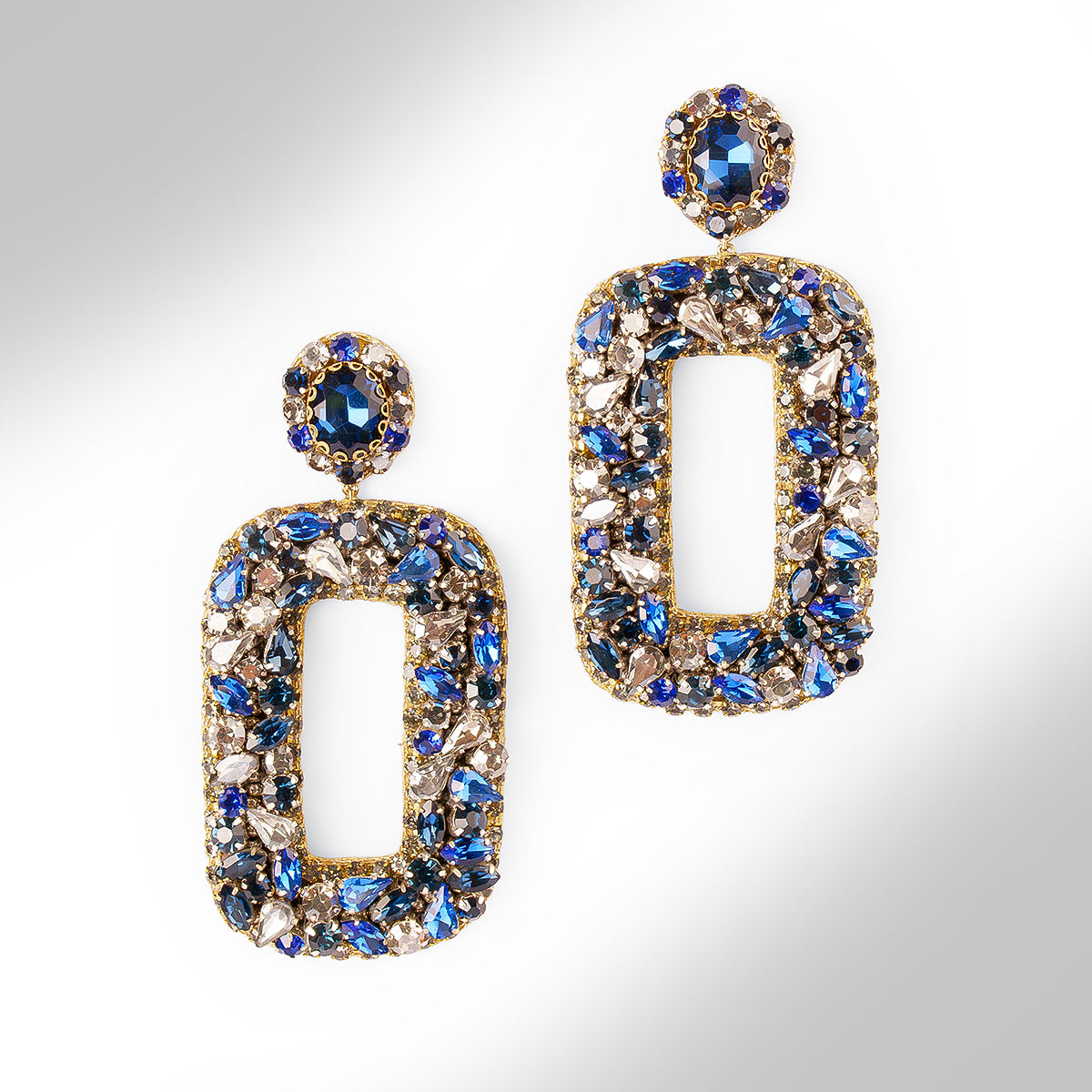 Gold-tone statement earrings with large open rectangular shapes and a matching round stud, heavily embellished with blue, silver, and clear faceted stones on a light gray background.