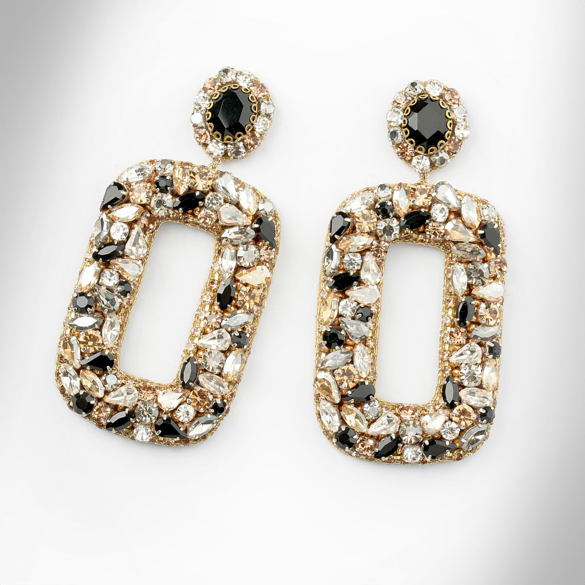Gold-tone statement earrings with large open rectangular shapes, heavily embellished with clear, white, and black faceted crystals shown on a light gray background.