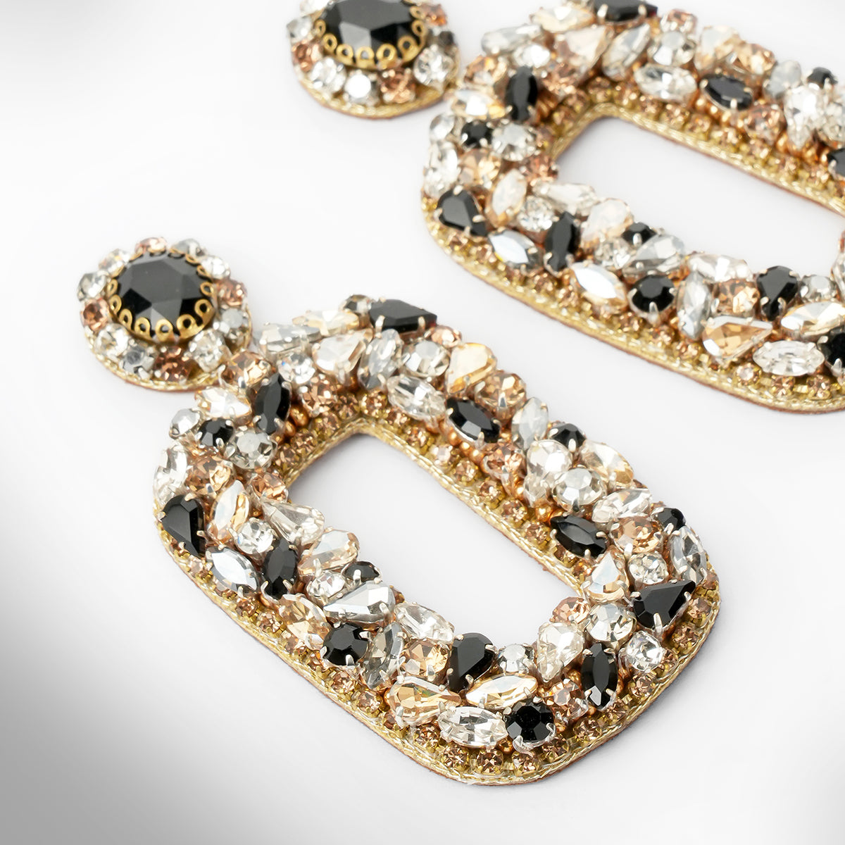 Close up of Gold-tone statement earrings with large open rectangular shapes, heavily embellished with clear, white, and black faceted crystals shown on a light gray background.