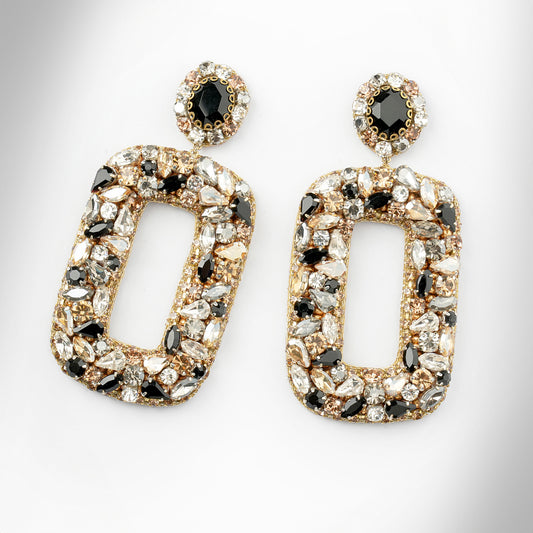 Gold-tone statement earrings with large open rectangular shapes, heavily embellished with clear, white, and black faceted crystals shown on a light gray background.