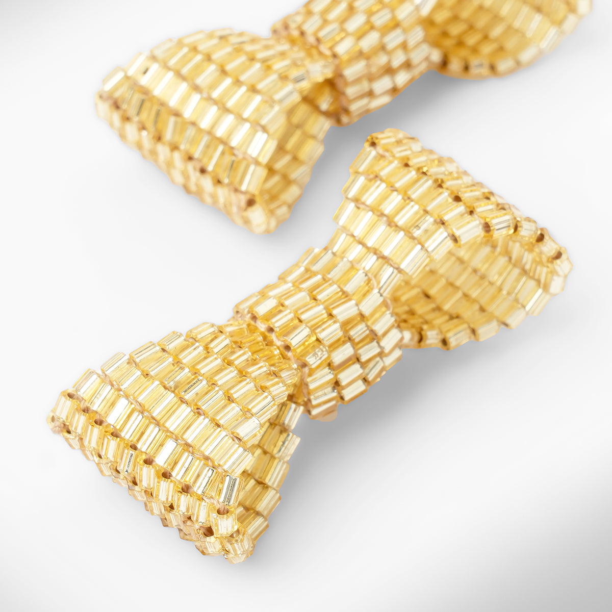 Close up of gold beaded bow-shaped earrings on a light gray gradient background.