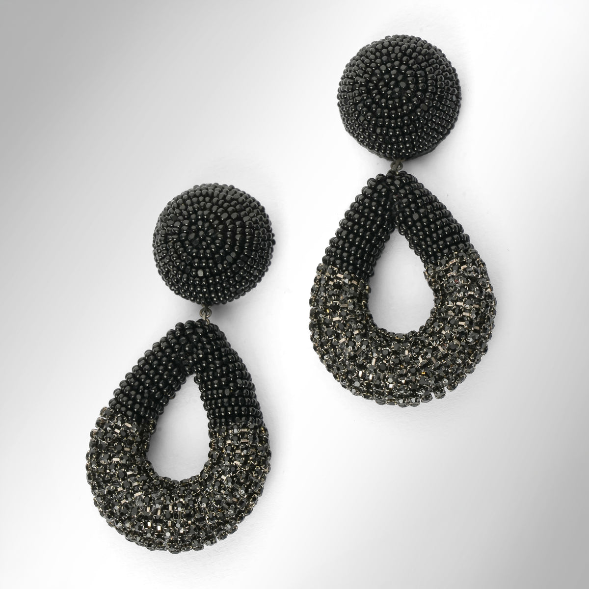 A pair of large statement dangle earrings made from black beads and chainstone, featuring a round stud top and a teardrop-shaped drop, shown on a light gray gradient background.