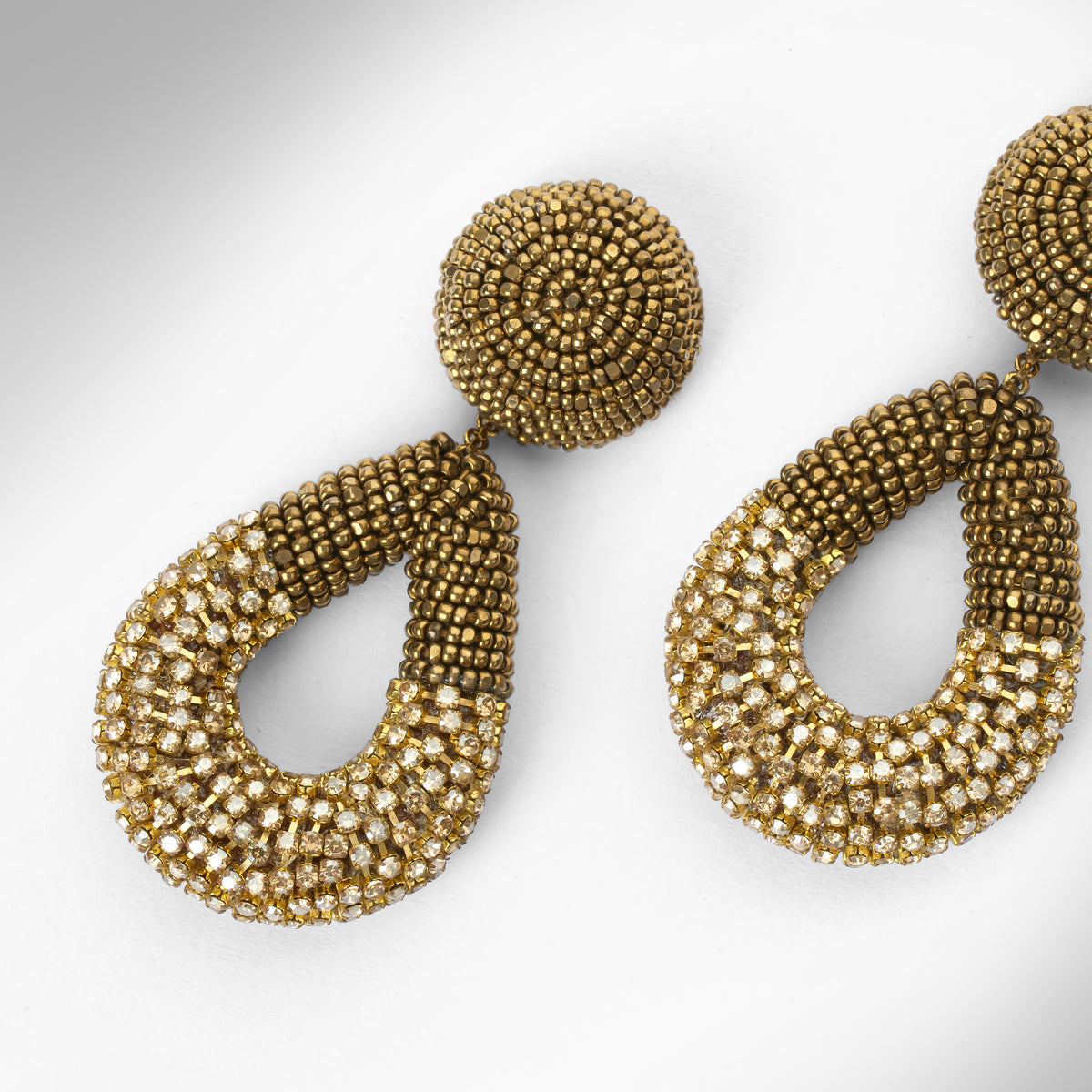 Close up of a pair of large statement dangle earrings made from gold tone beads and chainstone, featuring a round stud top and a teardrop-shaped drop, shown on a light gray gradient background.