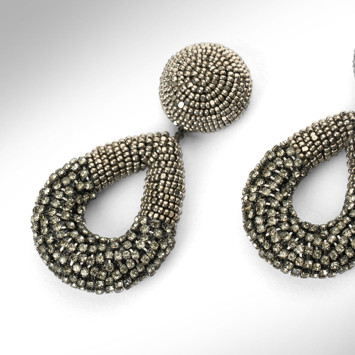 Close up of a pair of large statement dangle earrings made from gunmetal beads and chainstone, featuring a round stud top and a teardrop-shaped drop, shown on a light gray gradient background.