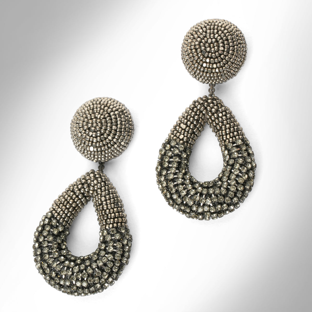 A pair of large statement dangle earrings made from gunmetal beads and chainstone, featuring a round stud top and a teardrop-shaped drop, shown on a light gray gradient background.