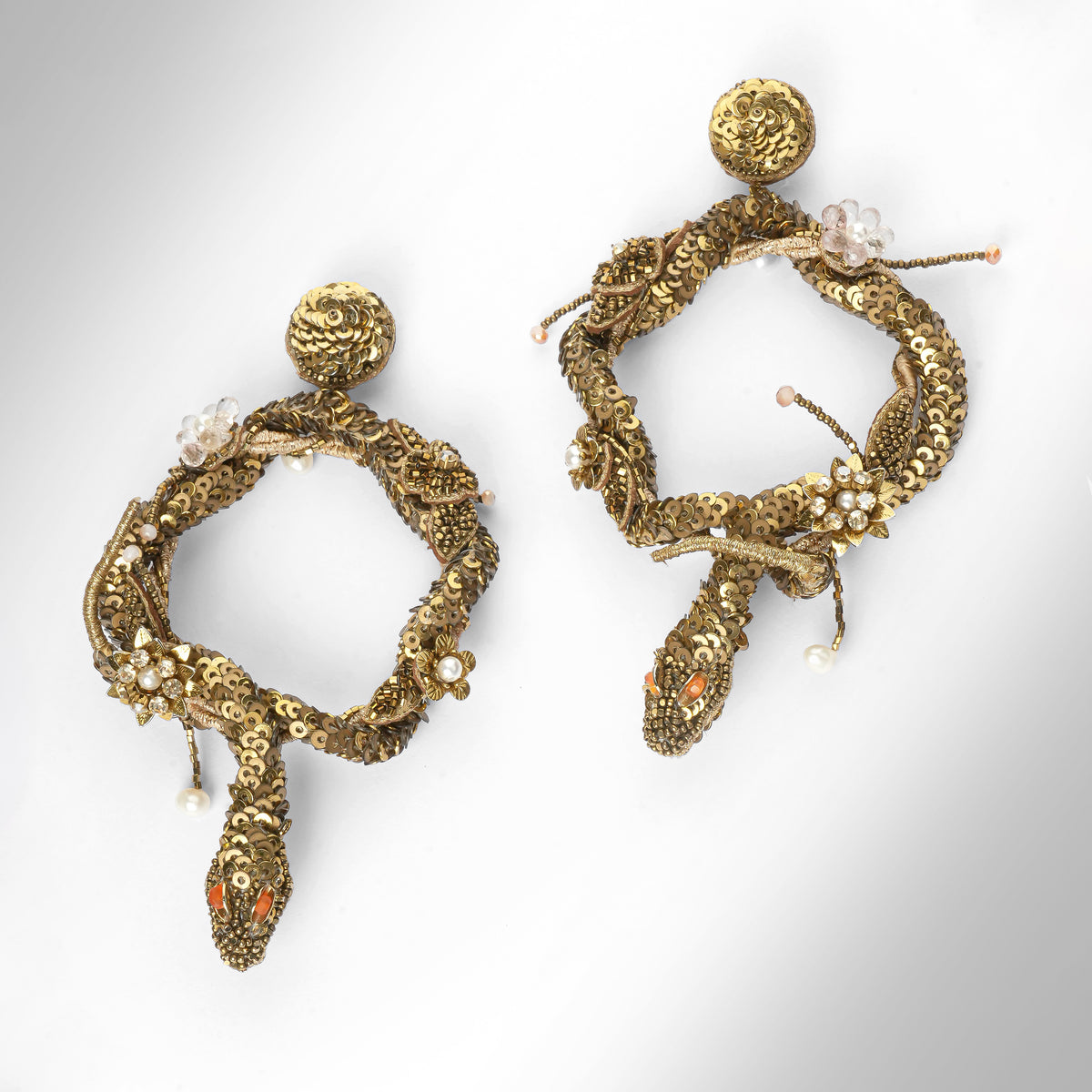 A pair of ornate gold-toned snake earrings with textured scales, small pearl-like beads, and orange-red eye details, photographed on a light gray background.