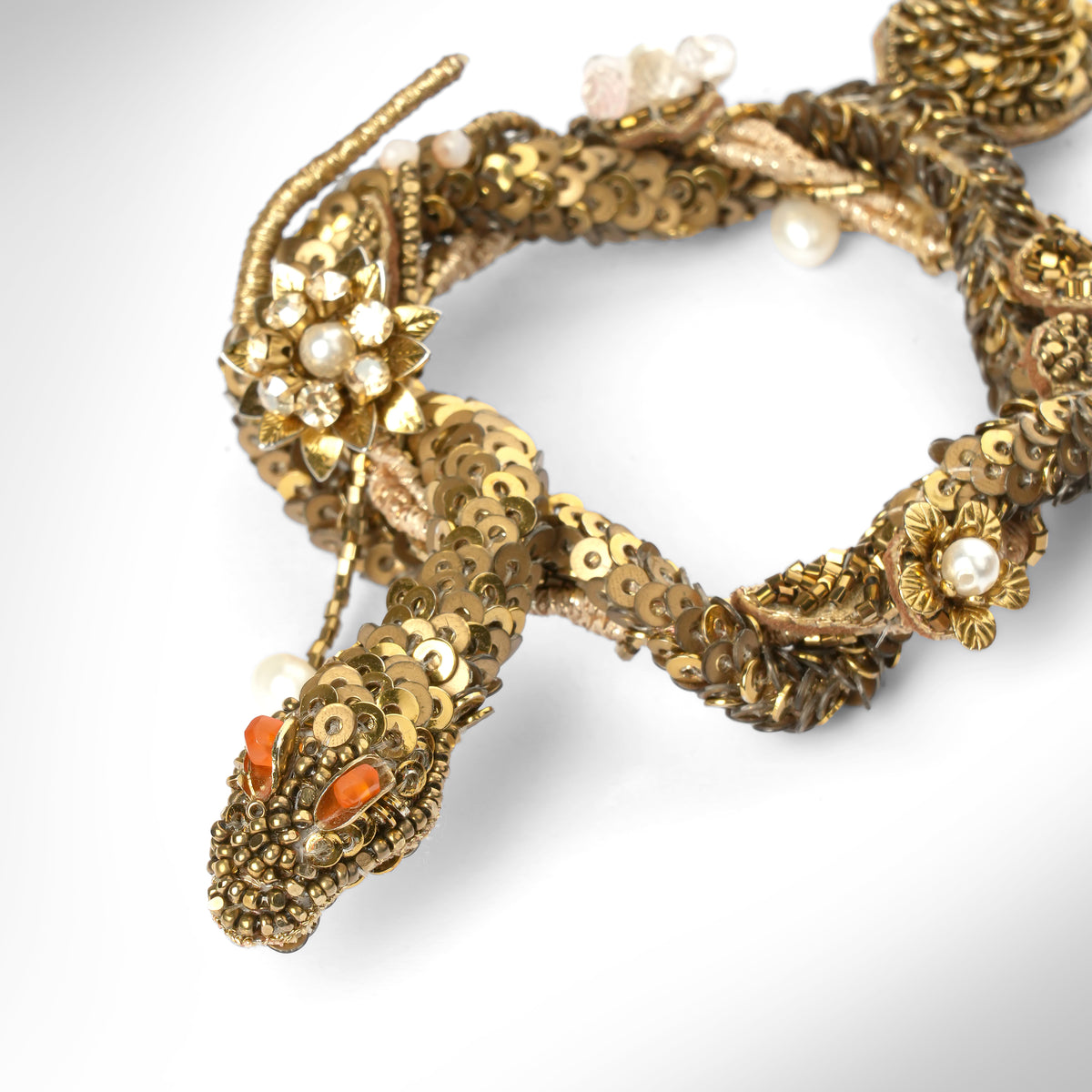 Close up of a pair of ornate gold-toned snake earrings with textured scales, small pearl-like beads, and orange-red eye details, photographed on a light gray background.