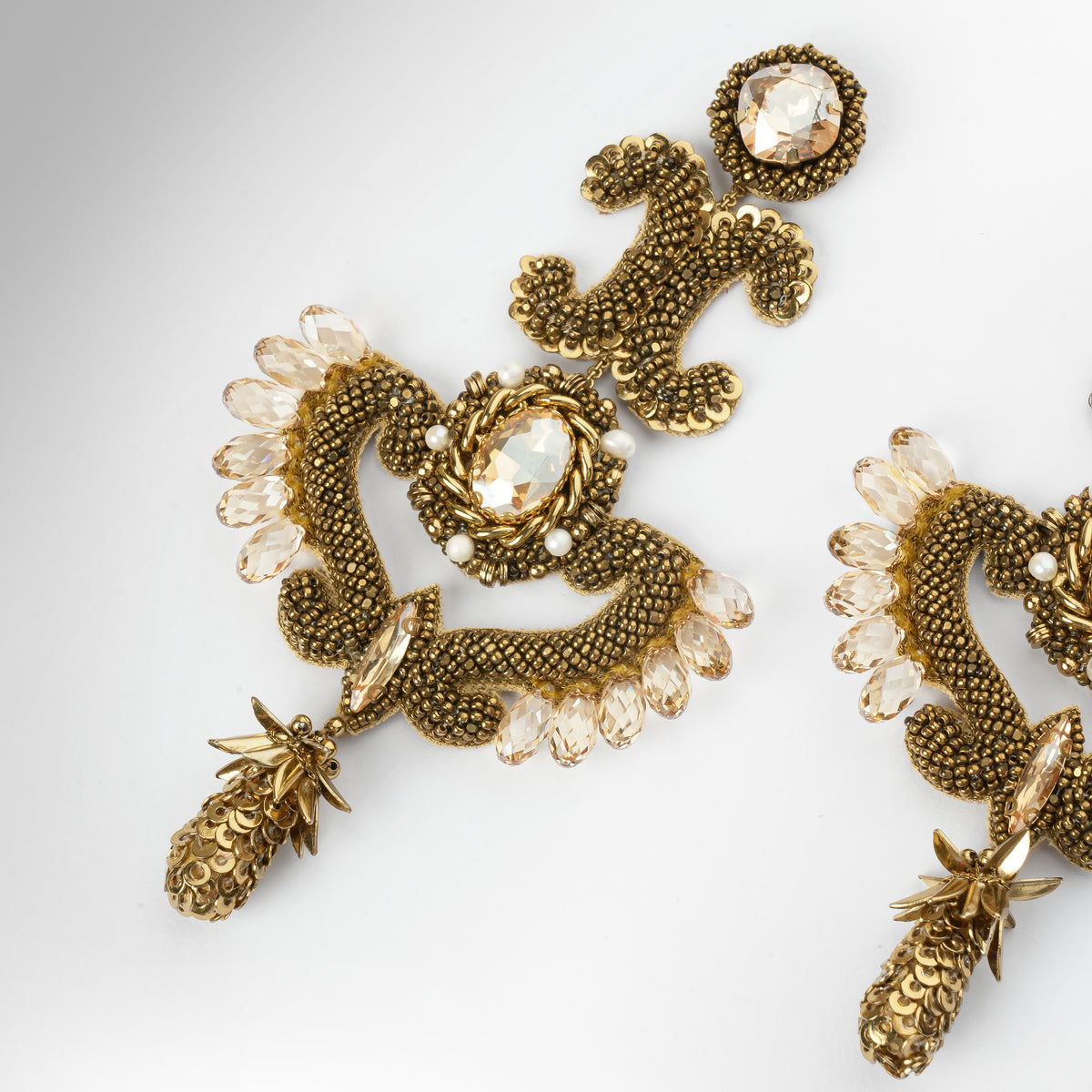 Close up of a pair of ornate gold-tone statement earrings with intricate beaded scrollwork and gold stone accents, displayed on a light gray background.