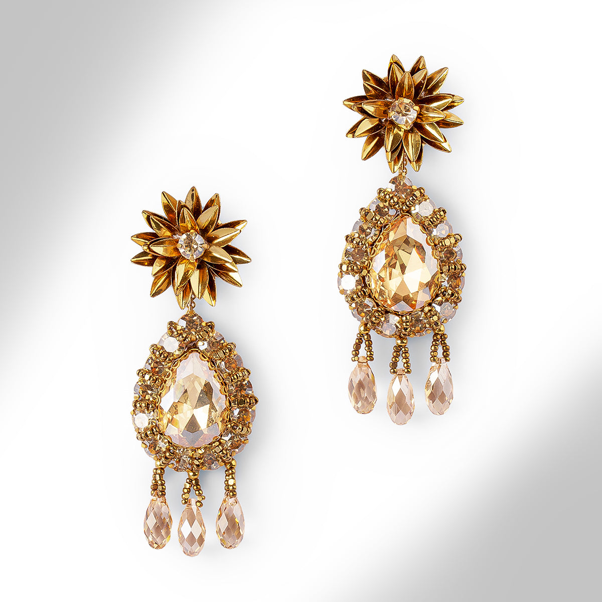 A pair of ornate gold-tone dangle earrings with starburst flower tops, teardrop-shaped crystals, and dangling teardrop beads, shown on a light gray gradient background.