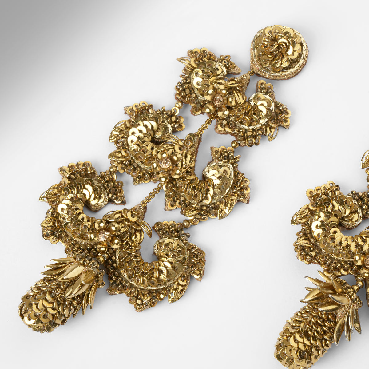 A close-up of an ornate gold-toned earring with intricate floral and leaf-like sculpted details, shown on a plain light gray background. 