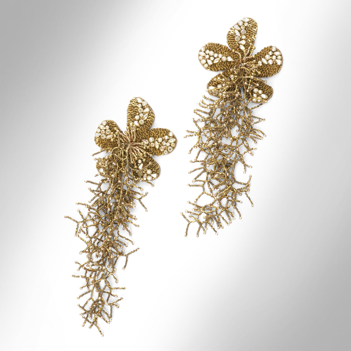 A pair of ornate gold statement earrings with floral tops and dangling  fringe, photographed on a light gray gradient background.