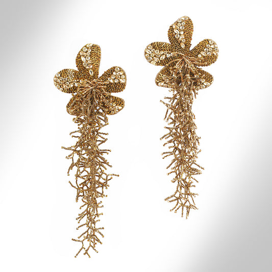 A pair of ornate gold statement earrings with floral tops and dangling  fringe, photographed on a light gray gradient background.