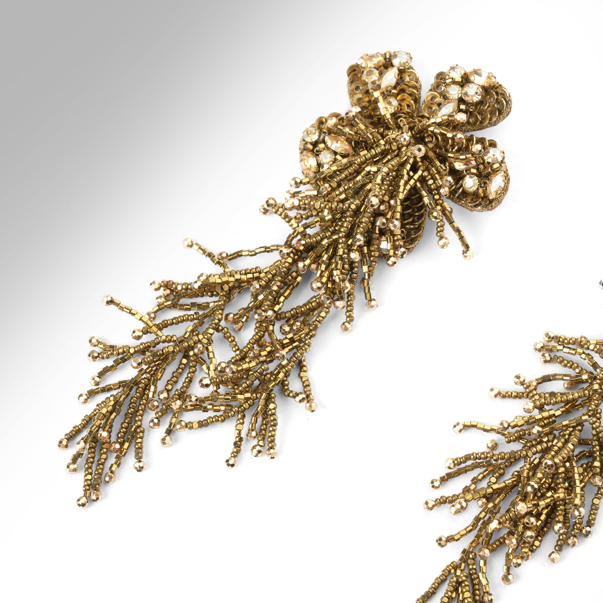 Close up of a pair of ornate gold-tone beaded earrings with a floral cluster at the top and cascading, branch-like beadwork hanging below, shown on a plain light background.