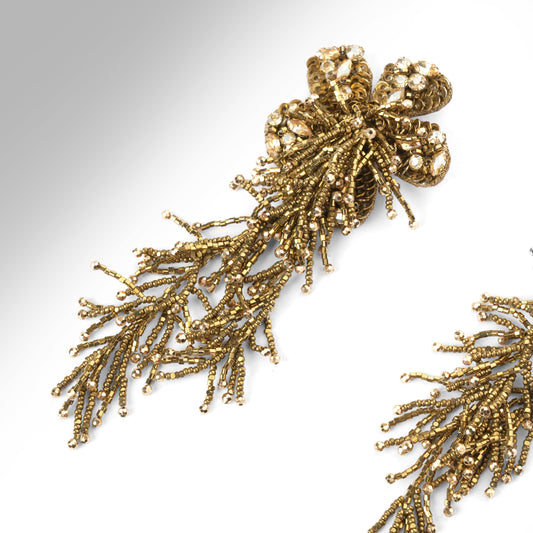 Close up of a pair of ornate gold-tone beaded earrings with a floral cluster at the top and cascading, branch-like beadwork hanging below, shown on a plain light background.