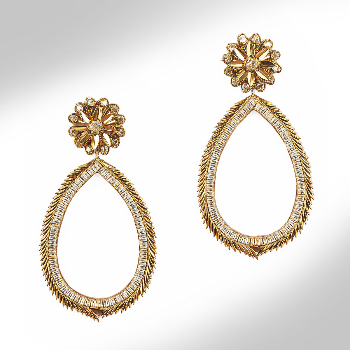 A pair of gold tone teardrop dangle earrings with crystal accents set against a light gray gradient background.