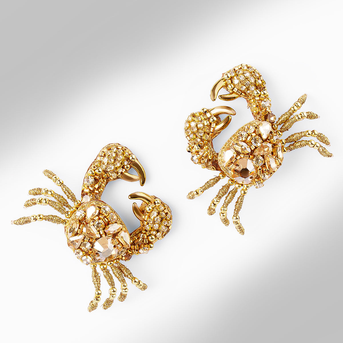 A pair of gold-toned crab-shaped earrings displayed on a light gray gradient background.