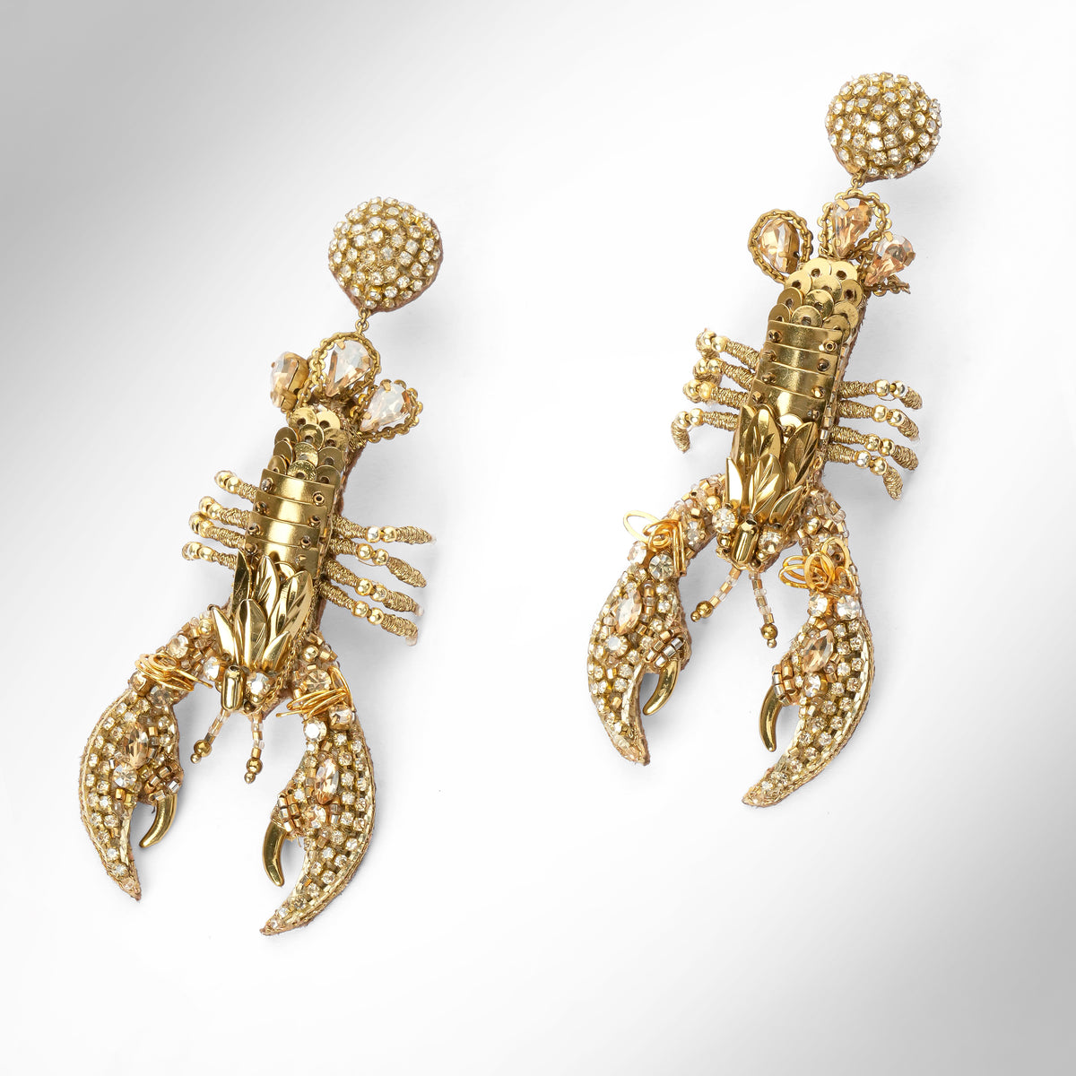 A pair of gold lobster-shaped drop earrings embellished with small gold stones, shown on a light gray gradient background.
